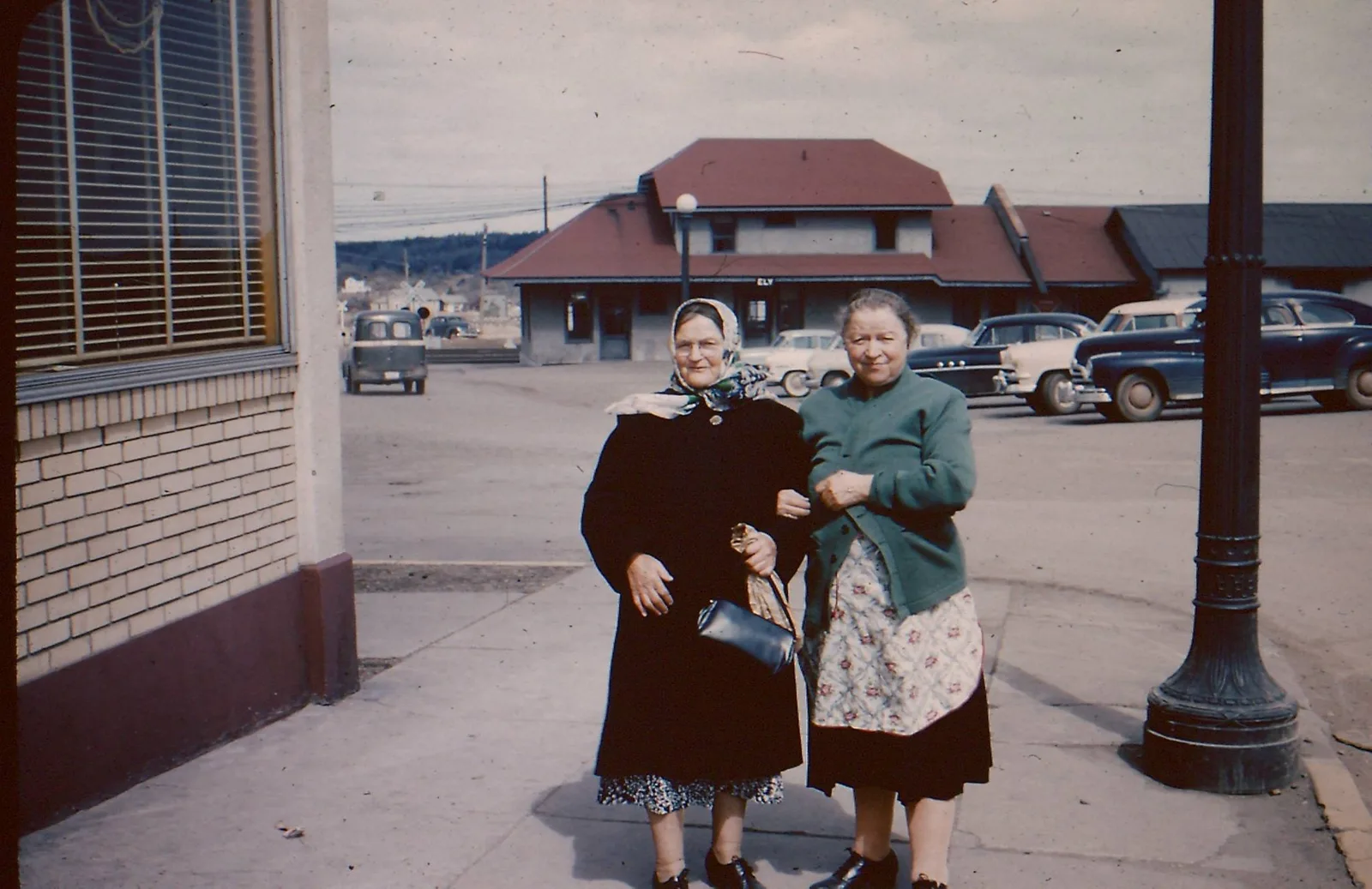 Two women posing on a sidewalk with the red-roofed depot behind them