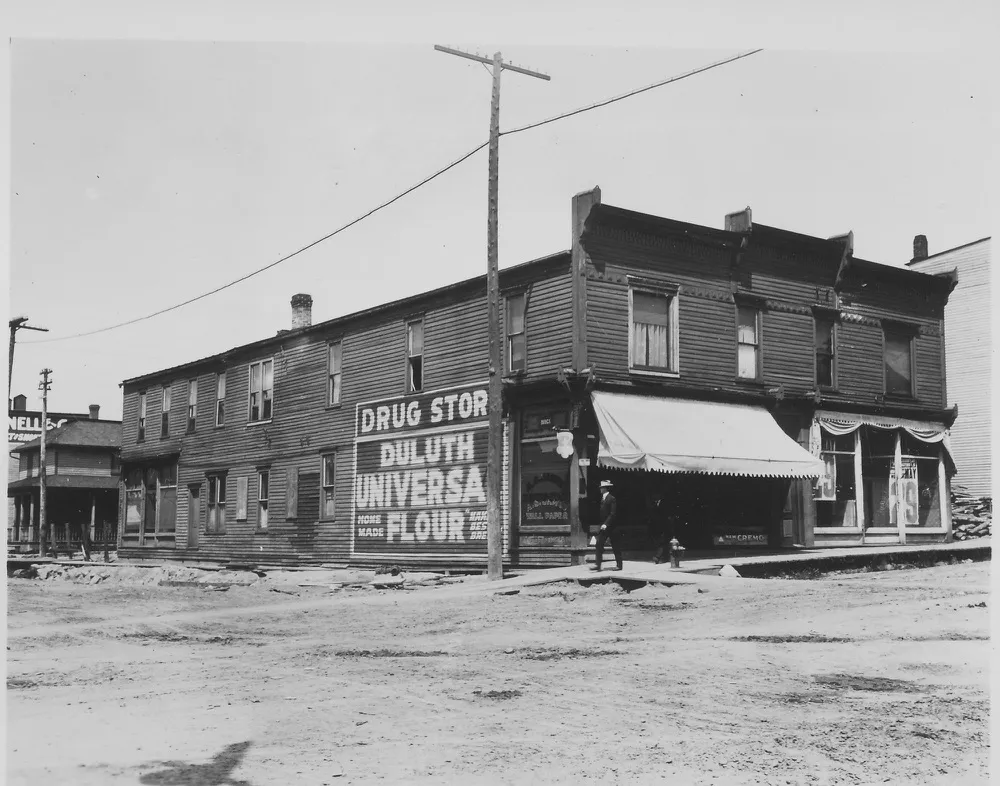 A two-story wood-frame drug store on a dirt street, its side wall painted Drug Store, Duluth Universal Flour