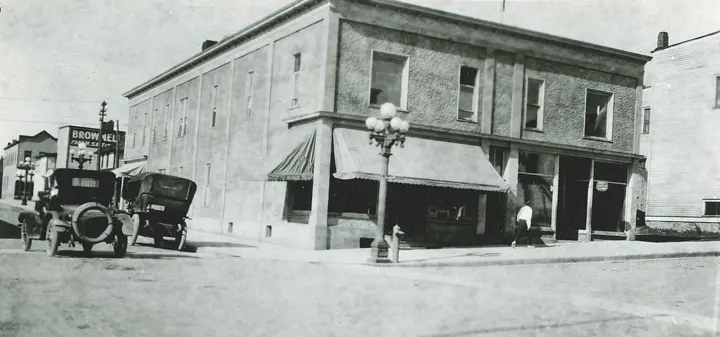 The two-story stucco building on the corner in its early years, with awnings, a globe street light, early automobiles and a Post Office sign in the window