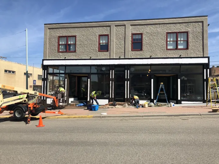 Crews installing new black storefront windows on the corner building with a lift parked at the curb