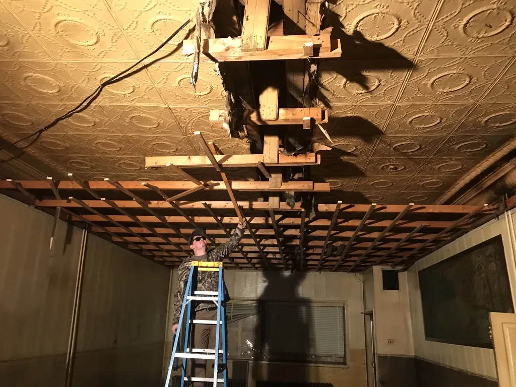 A worker on a ladder pulling down the drop ceiling framing to reveal a pressed tin ceiling