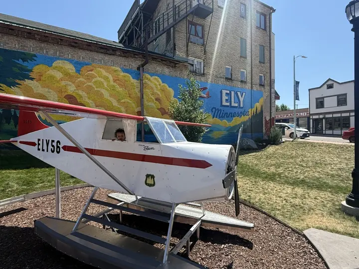A young child peeking out the window of the ELY56 floatplane in front of the Ely mural