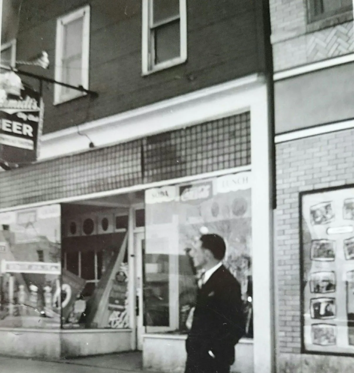 A man in a suit on the sidewalk in front of a storefront with Soda, Coca-Cola and Lunch signs, a glass block transom and a Schmidt Beer sign overhead