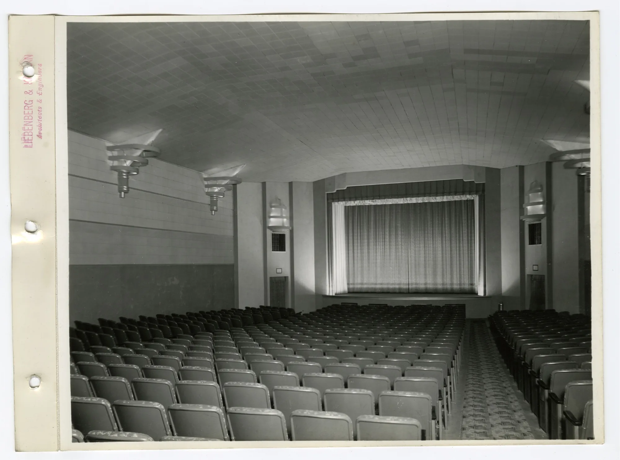 Theater auditorium with rows of seats, a curtained stage and tiered Art Deco wall and ceiling light fixtures