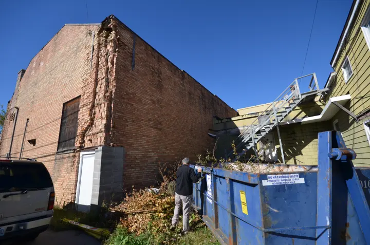 The back brick wall of the theater with a crumbling vertical section beside a dumpster