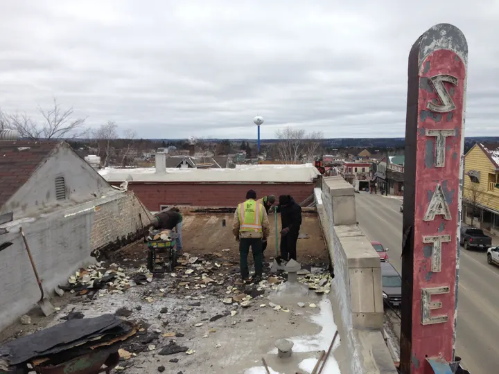 Roofers tearing off old roofing beside the STATE sign, with the Ely water tower in the distance