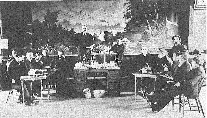 A lecture on the stage of the Ely Opera House with men seated around tables of scientific equipment