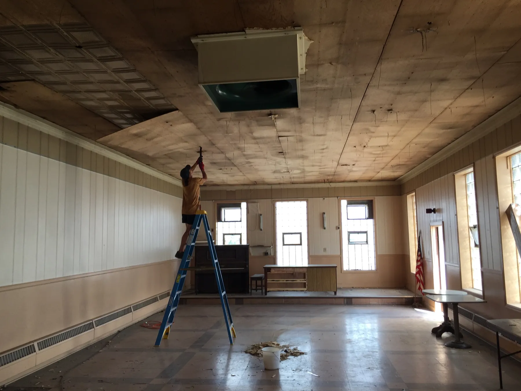 A worker on a ladder pulling plywood off the upstairs ceiling to reveal tin, with a piano and flag on the stage